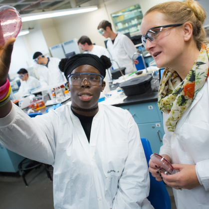 Two students in labcoats analyze petri dish. 