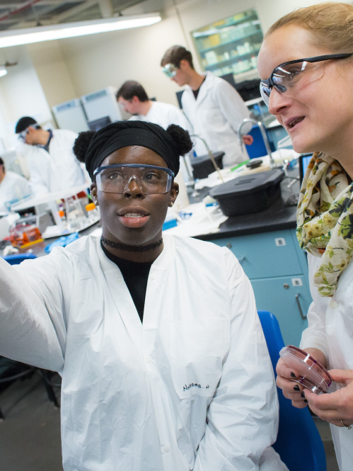 Two students in labcoats analyze petri dish. 